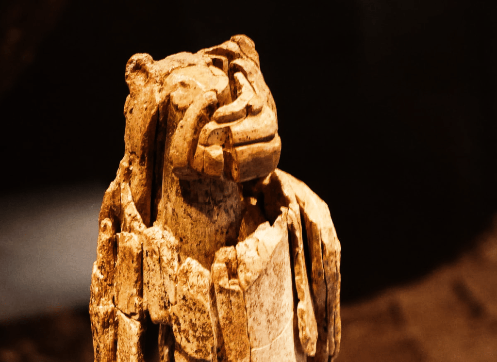 Escultura tallada en marfil de mamut que representa una figura antropomorfa con rasgos animales, destacada por una cabeza estilizada y un cuerpo vertical que muestra huellas del desgaste propio de su antigüedad. El trabajo minucioso en el rostro y en las líneas que recorren su superficie sugiere un simbolismo ritual o mitológico. Su estado fragmentado y la nobleza del material aportan un carácter histórico.